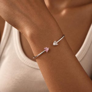 Op de afbeelding: Een zilverkleurige open armband met twee driehoekig geslepen stenen aan de uiteinden. De ene steen is een heldere, kleurloze edelsteen, terwijl de andere een lichtroze edelsteen is. De armband heeft een gladde, gepolijste metalen band.