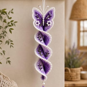 May include: A hanging crochet spiral ornament featuring a purple and white butterfly at the top. The spiral body is decorated with embroidered green leaves and small white flowers. A single purple crystal bead hangs from the bottom.