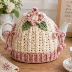 Puede incluir: Una funda de ganchillo para tetera en color crema con textura acanalada, una flor de ganchillo rosa con dos hojas verdes en la parte superior y una base acanalada rosa. La funda incluye lazos laterales rosas con pequeñas bolas de ganchillo.