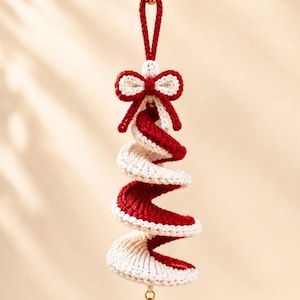 May include: A hanging spiral ornament crafted from red and white yarn, featuring a decorative bow at the top and a small gold-coloured metal bell at the bottom. The twisted, corkscrew-shaped design is suspended by a matching red yarn loop.