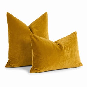 May include: Two mustard yellow velvet throw pillows. One pillow is square and positioned upright, while the other is rectangular and placed in front, slightly angled. Both pillows feature a soft, plush texture with a consistent solid color.