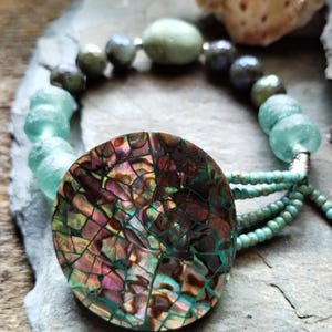 Puede incluir: Un collar artesanal con un gran colgante iridiscente con diseño de mosaico. El collar incorpora cuentas de vidrio verde mar, cuentas grises oscuras y un colgante central con un patrón de mosaico en tonos marrón, rosa y verde. Una pieza de joyería única.