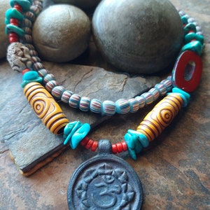 May include: A colourful necklace with turquoise, red and yellow beads, and a large, dark blue pendant with an Om symbol. The necklace is on a brown, textured surface.