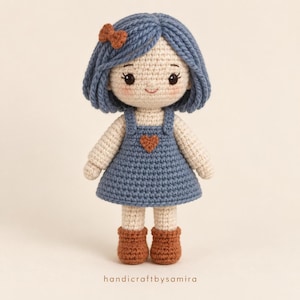 Könnte beinhalten: Eine gehäkelte Puppe mit blauem Wollhaar, einer kleinen braunen Schleife und einem blauen Kleid mit braunem Herz. Die Puppe hat ein cremefarbenes Gesicht mit schwarzen Augen und rosa Wangen sowie braune Stiefel. Unten steht der Text "handicraftbysamira".