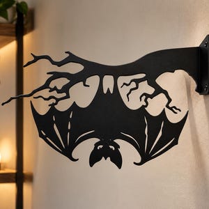 May include: A matte black metal wall-mounted bracket featuring a silhouette of a bat hanging upside down from a bare, gnarled tree branch. The design includes a flat rectangular mounting plate with two visible screw holes for attachment to a wall.