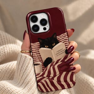 Könnte beinhalten: Eine dunkelrote Smartphone-Hülle mit einer Grafik eines schwarzen Katers, der ein Buch mit dem Titel COMMENT DOMESTIQUER LES HUMAINS liest, eingewickelt in eine rot-weiß gestreifte Decke neben einer kleinen Tasse. Die Hülle hat einen Dreifach-Kamera-Ausschnitt.