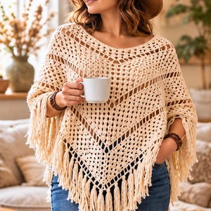May include: A cream-colored crochet poncho featuring an openwork diamond pattern and long fringe trim along the hem. The garment is worn with a wide-brimmed brown hat and blue denim jeans, while holding a plain white ceramic mug.