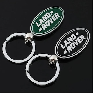 Könnte beinhalten: Zwei ovale Schlüsselanhänger aus Metall mit dem Land Rover-Logo. Einer hat einen dunkelgrünen Hintergrund mit weißem Text, der andere einen schwarzen Hintergrund mit weißem Text. Beide verfügen über einen silberfarbenen Schlüsselring.