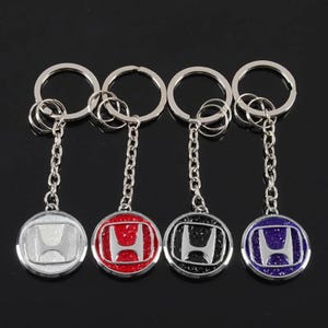 May include: Four metal keychains featuring the silver Honda logo emblem. Each keychain has a circular pendant with a textured background in white, red, black, or blue, attached to a silver chain and a circular split ring for holding keys.
