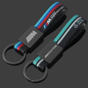 Könnte beinhalten: Zwei Schlüsselanhänger aus schwarzer Kohlefaser mit Metallringen. Einer zeigt das BMW M Logo und Schriftzug mit blauen und roten Streifen. Der andere zeigt das AMG Logo und das Branding des Mercedes-Benz Petronas Formula One Teams mit türkis-weißen Streifen.