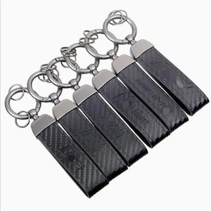Op de afbeelding: Een set van vijf rechthoekige sleutelhangers met een zwart koolstofvezelpatroon en metalen gunmetal hardware. Elke sleutelhanger bevat een ronde splitring en een rechthoekige band met in reliëf aangebrachte logo's, waaronder M, M-line en AMG.