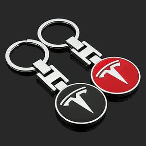 Pode incluir: Dois porta-chaves de metal com um pendente circular que apresenta o logótipo Tesla branco. Um pendente tem um fundo preto e o outro um fundo vermelho. Cada porta-chaves inclui um anel prateado ligado a um conector metálico retangular.