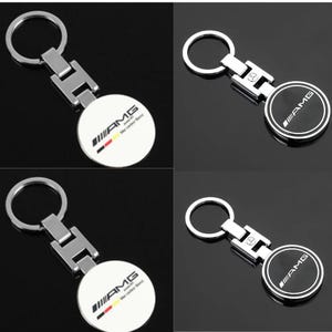 Könnte beinhalten: Ein Set aus vier Metallschlüsselanhängern mit einem runden Anhänger und einem silberfarbenen Ring. Zwei Schlüsselanhänger haben eine weiße runde Fläche mit dem schwarzen AMG-Logo und einer Deutschlandflagge. Zwei haben eine schwarze Fläche mit AMG-Logo.