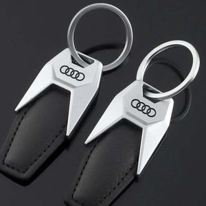 May include: Two silver-toned metal keychains featuring a black leather-like insert and the Audi logo engraved in black. Each keychain has a geometric, angular design and a circular split ring for keys, presenting a sleek and modern appearance.