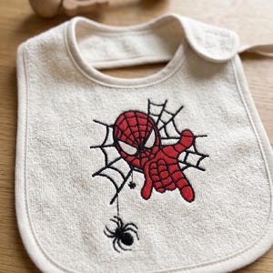 Peut inclure: Un bavoir pour bébé en tissu éponge blanc cassé avec une broderie représentant un super-héros rouge et noir dans une toile d'araignée, accompagnée d'une petite araignée noire suspendue. Le bavoir a une encolure arrondie.