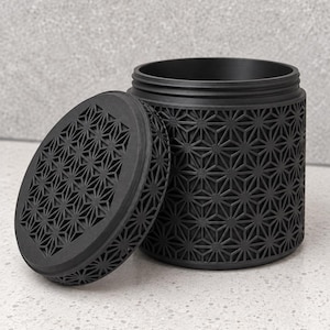 Pode incluir: Um recipiente cilíndrico preto fosco com uma tampa correspondente, ambos apresentando um padrão geométrico de estrelas em relevo em suas superfícies. O recipiente está aberto, revelando um interior oco, e a tampa está ao lado.
