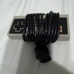 Pode incluir: Um comando de videojogos retangular cinzento com um pad direcional preto à esquerda, dois botões circulares vermelhos à direita e um painel central preto. Um cabo preto comprido e enrolado com um conector retangular envolve o comando.
