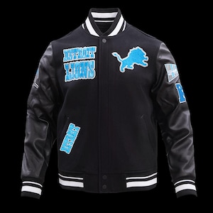 May include: A black varsity jacket with black leather sleeves, featuring the text Detroit Lions in blue, a blue lion logo, and white and black striped ribbed trim on the collar, cuffs, and hem. The jacket includes additional blue patches.