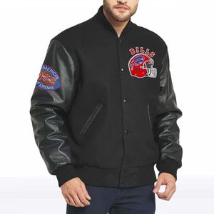Puede incluir: Una chaqueta universitaria negra con mangas largas negras, cierre frontal de botones a presión y puños acanalados. El pecho muestra un logotipo de casco rojo de los Buffalo Bills con el texto BILLS, y la manga tiene un parche de la AFC.