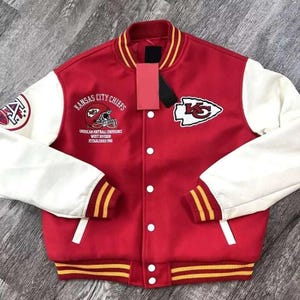 Pode incluir: Uma jaqueta universitária vermelha e branca com o logotipo dos Kansas City Chiefs, um gráfico de capacete de futebol americano e o texto American Football Conference, West Division, Established 1960. Possui mangas brancas, punhos canelados vermelhos com listras amarelas e botões brancos.