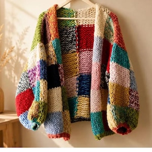 Op de afbeelding: Een grof gebreid vest met een patchworkontwerp van grote vierkanten in diverse kleuren, waaronder rood, blauw, geel, groen en crème. Het kledingstuk heeft lange mouwen met geribbelde boorden en een open voorzijde.