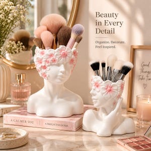 Peut inclure: Deux organisateurs blancs en forme de tête avec des accents floraux roses contenant des pinceaux de maquillage. Le plus grand présente une couronne florale, le plus petit montre une figure avec une main près du visage. Texte Beauty in Every Detail.