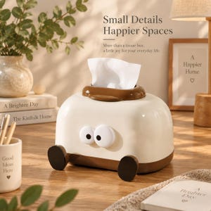 May include: A cream-coloured tissue box cover shaped like a toaster with a brown base and top rim. It features two large white eyes with black pupils on the front and two small brown feet at the bottom. A white tissue is pulled through the top slot.