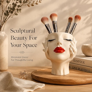 Peut inclure: Un porte-pinceaux en forme de tête couleur crème avec des mains encadrant le visage, des yeux fermés et des lèvres rouge vif. Quatre pinceaux de maquillage aux poils bruns sont insérés au sommet. Le texte indique : Sculptural Beauty For Your Space, Minimalist Decor For Thoughtful Living.