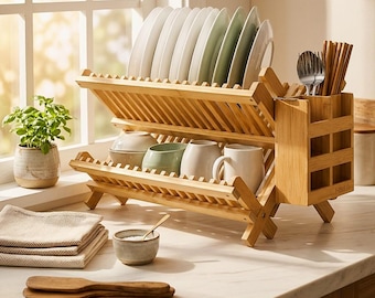 Pode incluir: Um escorredor de pratos de bambu de dois níveis com um suporte lateral para utensílios. O nível superior contém vários pratos brancos, enquanto o nível inferior acomoda tigelas e canecas. O suporte é feito de ripas de madeira clara.