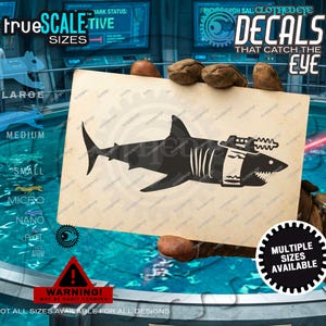 May include: A rectangular card featuring a black silhouette of a shark with a laser cannon mounted on its back. The shark has a white band around its midsection. The card has a light tan, textured background and is held by gloved hands.