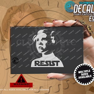 May include: A black decal featuring a white graphic of a woman's face with the word "RESIST" below. The decal is held in a hand. Text on the decal reads "CLOTHED EYE DECALS THAT CATCH THE EYE". A warning sign and a "MULTIPLE SIZES AVAILABLE" sticker are also present.