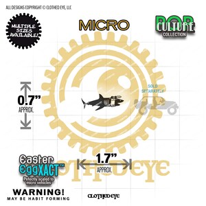 May include: A black shark silhouette sticker with a mechanical attachment, featuring text including MICRO, POP CULTURE COLLECTION, and Easter EggXACT. The design includes dimension markers indicating approximately 0.7 inches in height and 1.7 inches in width.