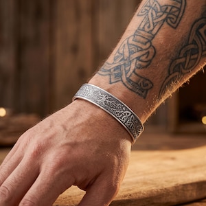 May include: A silver-toned metal cuff bracelet featuring an intricate embossed design of a tree with sprawling branches and interwoven Celtic knotwork patterns along the entire length of the band, showcasing a dark, oxidized finish in the recessed areas.