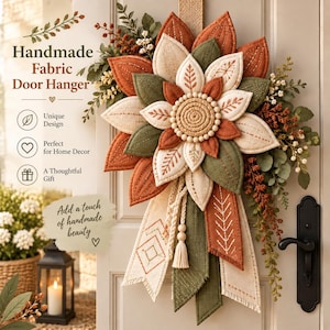 Könnte beinhalten: Ein Türhänger aus Stoff mit einem mehrschichtigen Blumendesign aus cremefarbenen, olivgrünen und rostfarbenen Blütenblättern. Die Mitte besteht aus einem strukturierten Seilkreis mit Holzperlen. Lange Stoffbänder mit Stickmustern hängen herab.