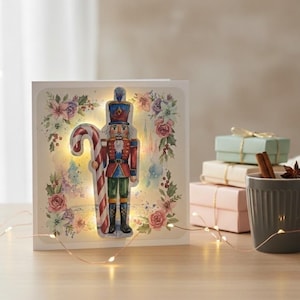 Könnte beinhalten: Eine quadratische Grußkarte mit einer zentralen Illustration eines Nussknackers, der eine große Zuckerstange hält, umgeben von einem Blumenkranz aus Rosen und Stechpalmen. Die Karte ist mit kleinen warmen LED-Lichtern beleuchtet.