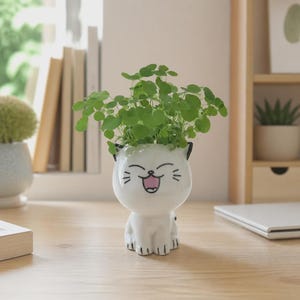 Puede incluir: Una maceta de cerámica blanca con forma de gato sentado, con cara sonriente, ojos cerrados y detalles pintados en negro. La figura del gato tiene una amplia abertura en la parte superior que contiene una pequeña planta verde de hojas redondas.