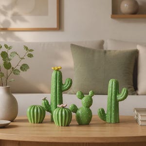 Op de afbeelding: Een verzameling van vier groene keramische cactusbeeldjes in verschillende vormen en maten. Twee hoge zuilvormige cactussen hebben kleine gele en roze bloemen aan de bovenkant, terwijl twee kortere, ronde cactussen gestructureerde oppervlakken met witte stippen hebben.