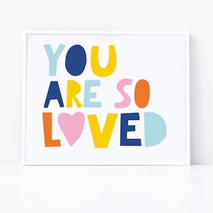 以下が含まれることがあります： 遊び心のあるフォントで「YOU ARE SO LOVED」というカラフルなプリント。文字は青、黄、オレンジ、ピンク、水色など、さまざまな色で書かれています。デザインにはピンクのハートが含まれています。