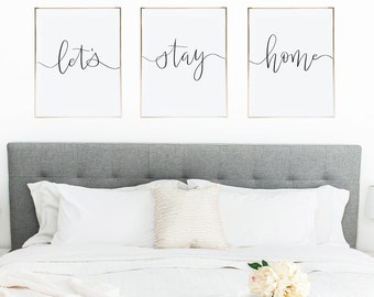 May include: Three framed prints with gold frames hanging on a white wall above a gray headboard. The prints feature the words "let's", "stay", and "home" in black cursive script.