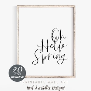 May include: Printable wall art featuring a black and white calligraphy design that says "Oh Hello Spring". The design is framed in a light wood frame.