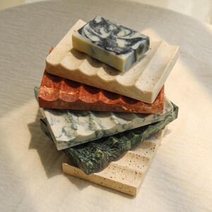 Peut inclure: Une pile de cinq porte-savons rectangulaires avec une surface ondulée et rainurée. Les porte-savons sont fabriqués en diverses pierres de couleurs beige, rouge, blanche et vert foncé. Un savon rectangulaire au motif marbré noir et blanc est posé dessus.
