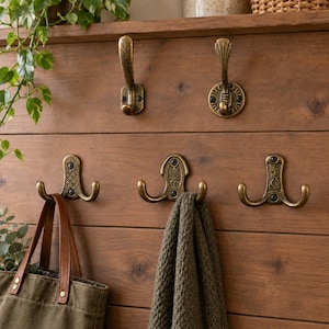 Op de afbeelding: Vijf wandhaken van metaal met een antieke messing afwerking, gemonteerd op een horizontale houten wand. De haken hebben diverse ontwerpen, waaronder stijlen met één of twee haken en decoratieve patronen, waaraan een groene tas en een grijze sjaal hangen.