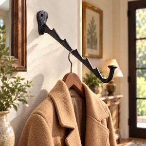 Puede incluir: Un perchero de pared de metal negro con un brazo diagonal que presenta varias muescas para colgar objetos. Un abrigo de lana color canela cuelga de una percha de madera suspendida en una de las muescas del soporte fijado a la pared.