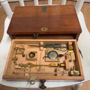 Könnte beinhalten: Ein hölzerner Instrumentenkoffer mit Klappdeckel und einem separaten Einsatz, der verschiedene Messingkomponenten enthält, darunter kleine Stäbe, eine kreisförmige Metallschale, einen Pinsel und Gelenkhebel, die in passenden Holzfächern angeordnet sind.