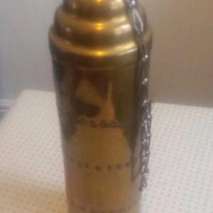May include: A cylindrical brass container with a rounded, tiered top and a metallic chain attached to the side. The surface of the brass object features horizontal bands of decorative etched patterns encircling the body of the vessel.