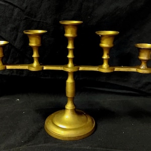 May include: A gold-toned metal candelabra featuring five candle holders arranged in a horizontal row. The central holder is elevated on a vertical stem supported by a circular base, with two additional holders extending on each side.