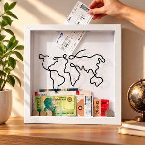 May include: A white shadow box frame featuring a black continuous line drawing of a world map on the glass. Inside the frame are various travel mementos including a 1000 Albanian Lek banknote, airline boarding passes, and metal coins.