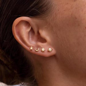 May include: A set of four gold stud earrings worn in a row on an earlobe. The earrings feature celestial designs including a five-pointed star, a crescent moon, a sun with a central stone, and a small planet with a ring around its center.
