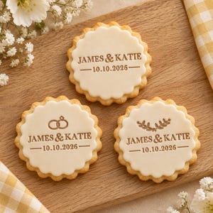 Puede incluir: Tres galletas redondas de borde festoneado cubiertas con glaseado blanco liso. Cada galleta presenta el texto JAMES & KATIE y la fecha 10.10.2026 en relieve marrón. Una galleta muestra dos anillos entrelazados, otra una rama.