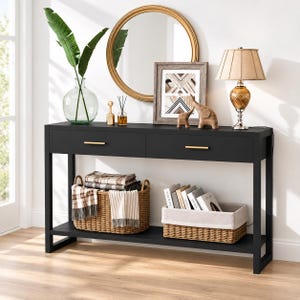 May include: A black console table with two drawers featuring gold handles, a lower shelf holding a woven basket with plaid blankets and a second woven basket with books, topped with a round mirror, a lamp, a picture frame, and a vase.